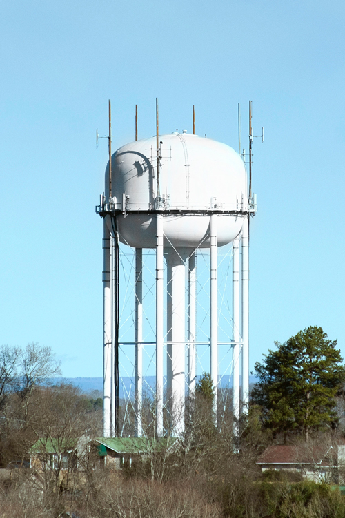 Tall white elevated water tower located on Highway 58 with support columns and antennas, rising above trees and nearby buildings